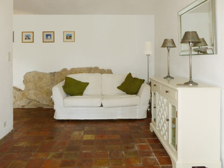 Moulin main living room on lower ground floor viewed through the french windows leading to the tearrace. The floor is tiled and the room painted white with photographs of windmills on the wall. There is a white sofa and white sideboard with a mirror above.